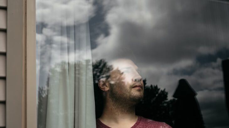 Thoughtful woman looks out a window at a dark, stormy sky, symbolizing the dread of a migraine triggered by changing barom...