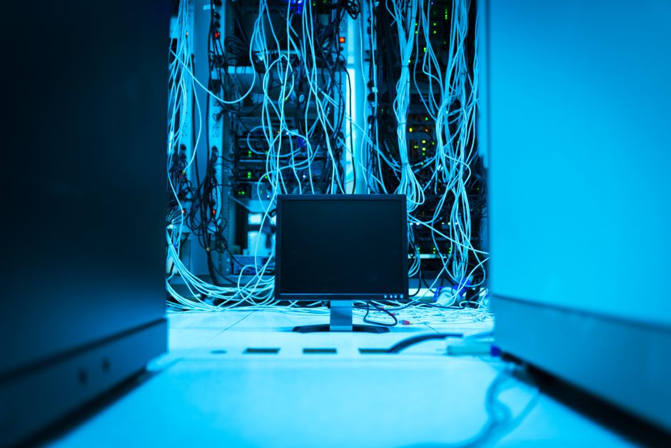 A lone professional standing in a server room, dwarfed by a massive and complex wall of servers and tangled cables, illustrating the overwhelming nature of large-scale enterprise technology.